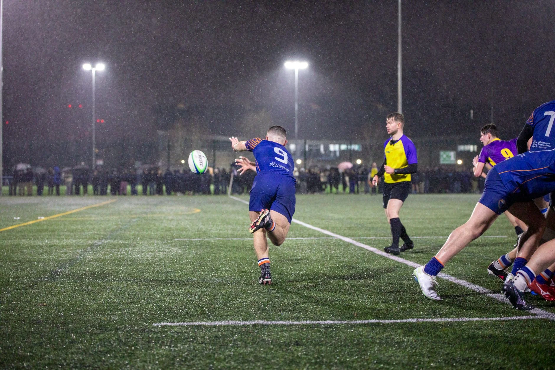 A rugby player catching the ball during a rain-soaked night match under floodlights