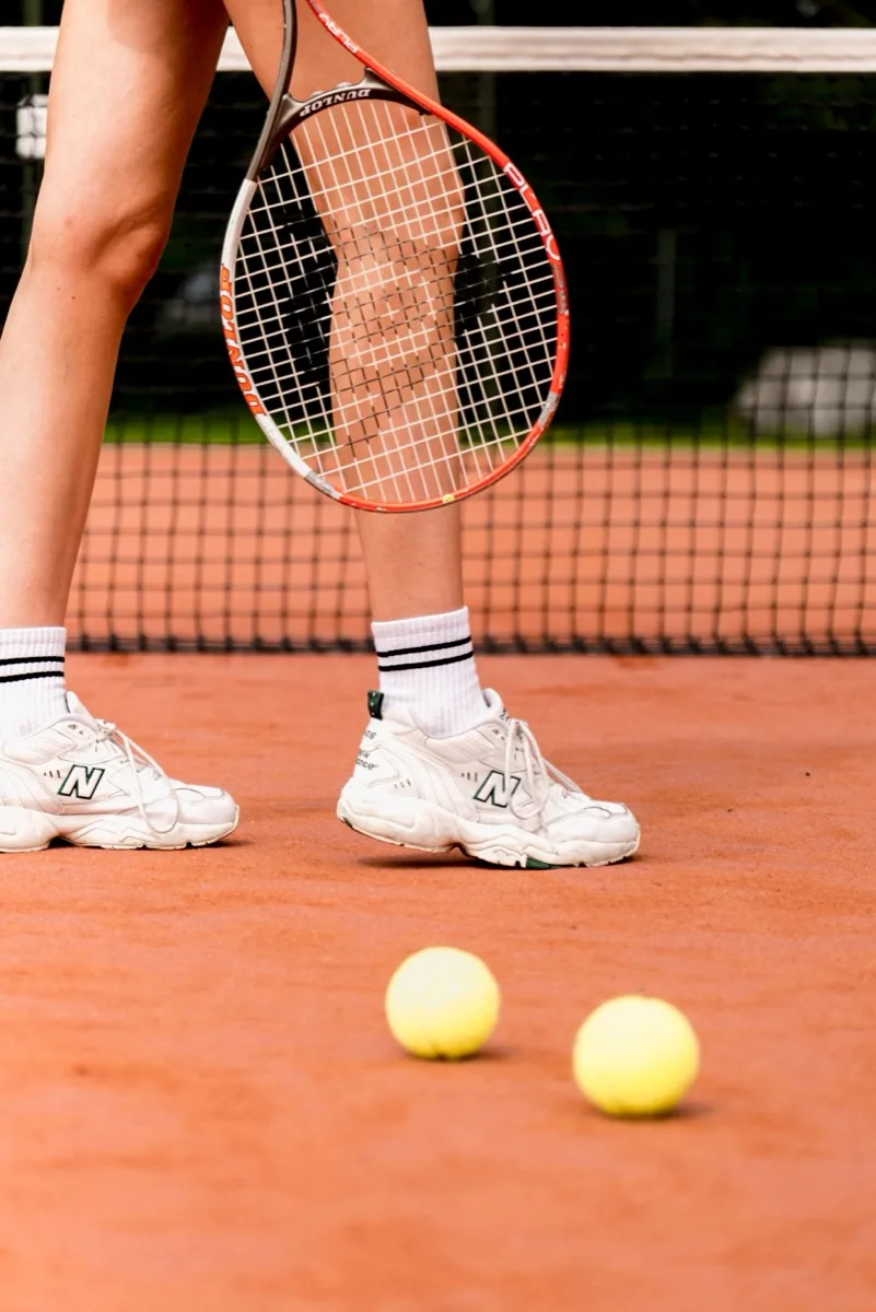 A player's sneakers beside two tennis balls on a clay court