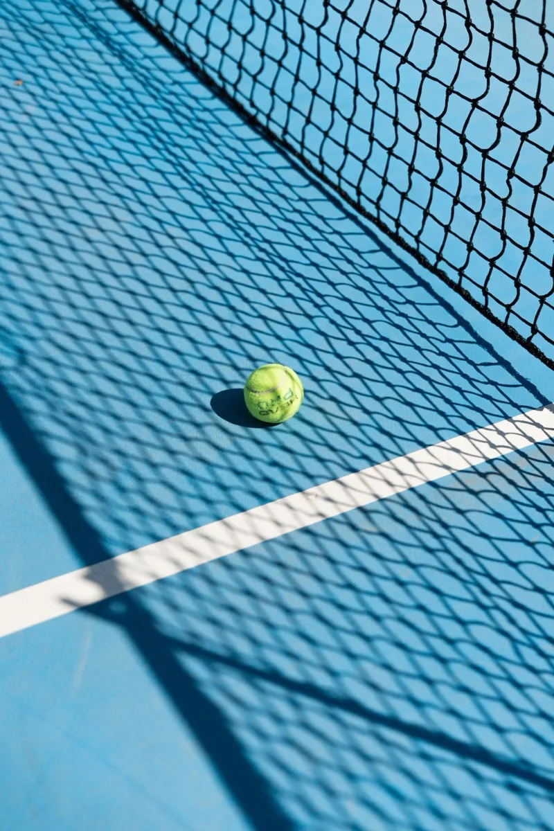 A tennis ball on a blue hard court beside the net shadow