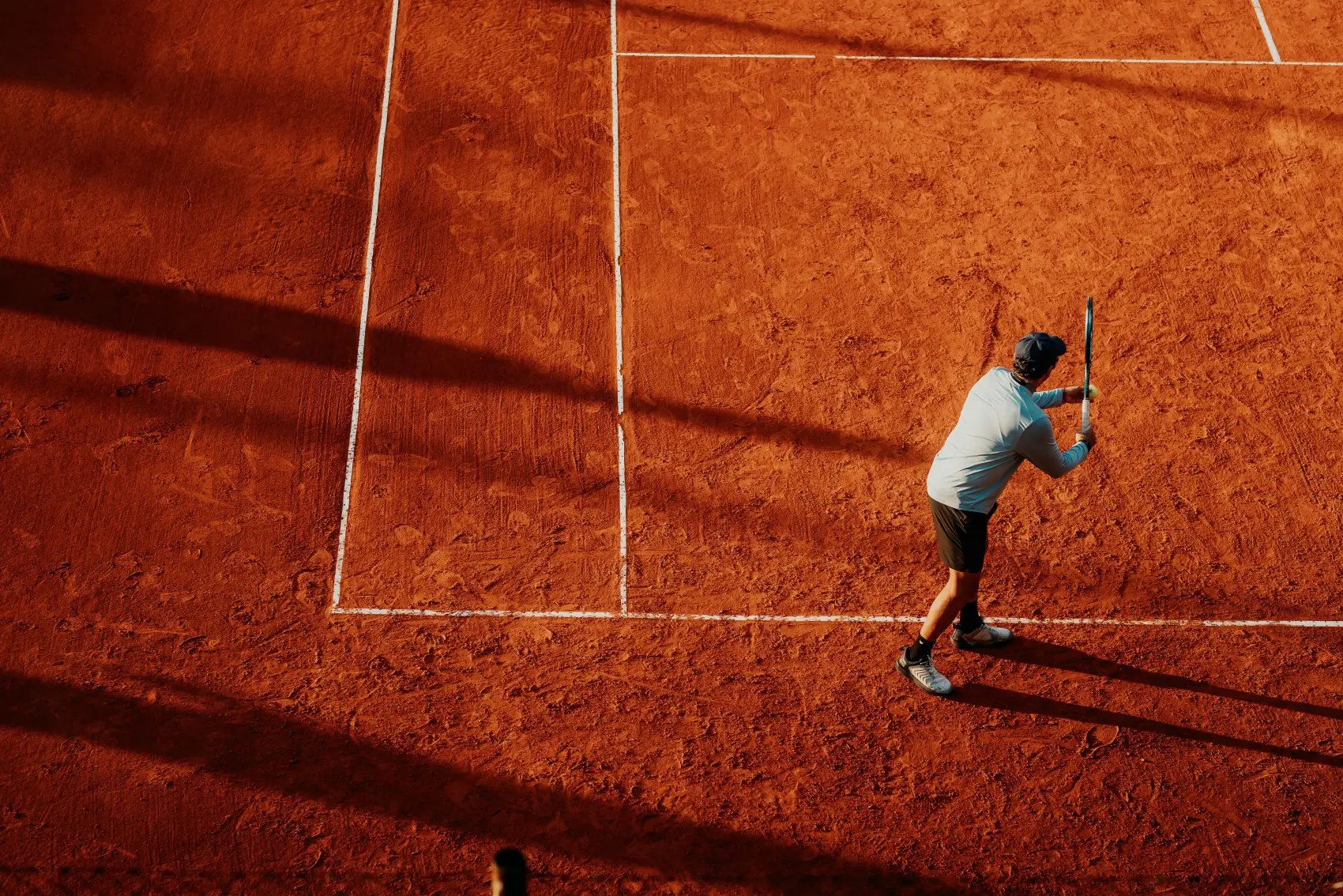 Aerial view of a tennis player serving on a clay court in dramatic afternoon light