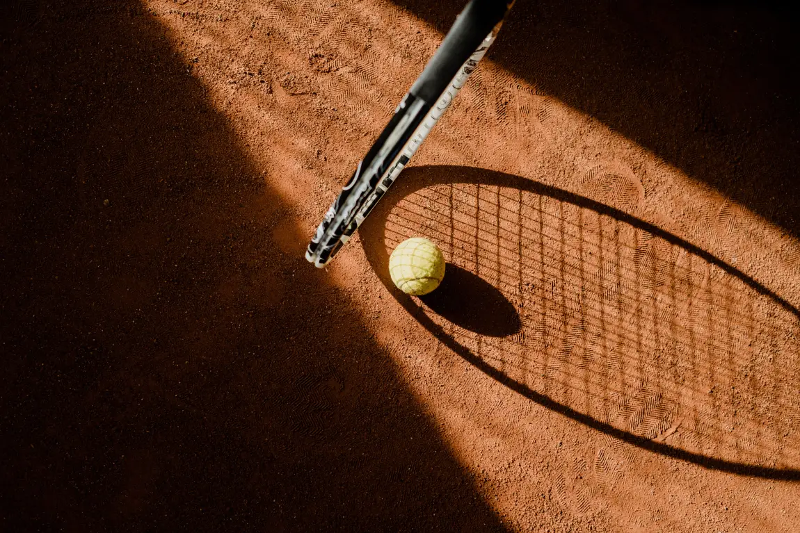 Close-up of a tennis racket and ball casting a long shadow on a clay court